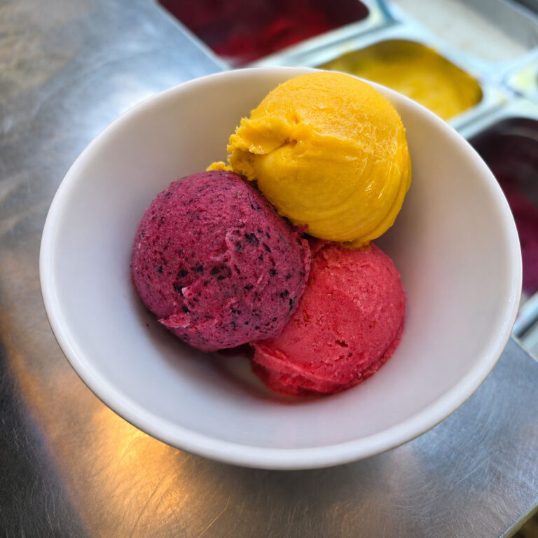 Drei farbenfrohe Sorbetkugeln in Gelb, Rot und Violett, frisch angerichtet in einer weißen Schale.