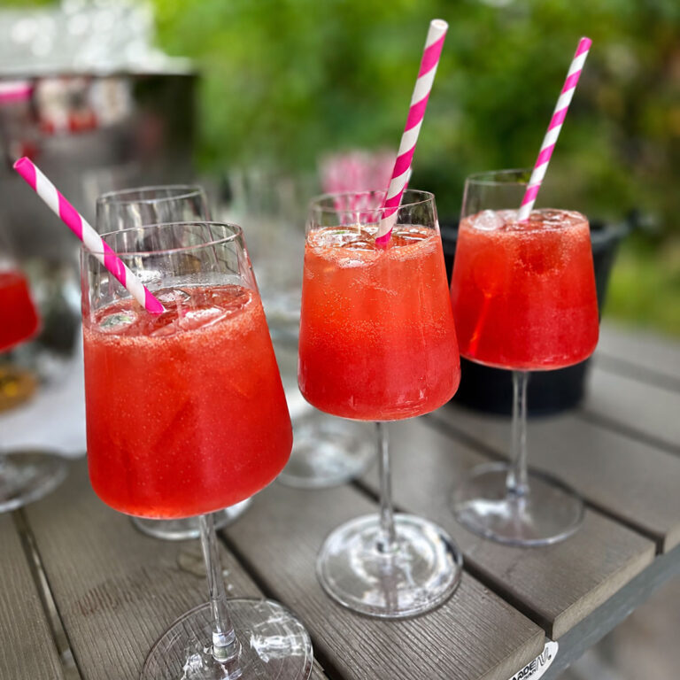 Drei leuchtend rote, eisgekühlte Sommerdrinks mit Strohhalmen, frisch serviert auf einer Terrasse mit grüner Biergarten-Kulisse.