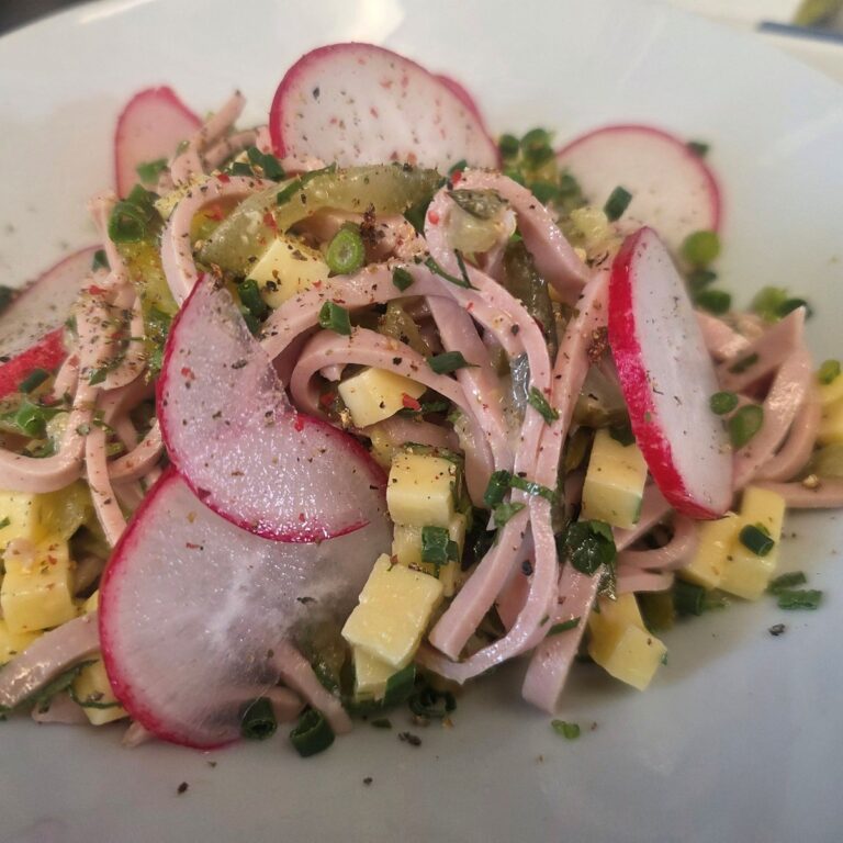 Frisch angerichteter Wurstsalat mit Käsewürfeln, Radieschenscheiben, Gewürzgurken und Schnittlauch, abgeschmeckt mit Pfeffer und Kräutern.